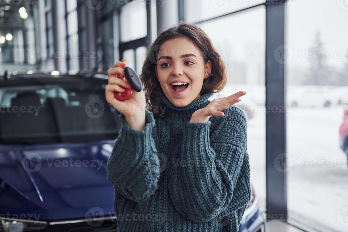 Fröhliche junge Frau hält Schlüssel und steht drinnen vor einem modernen neuen Auto foto