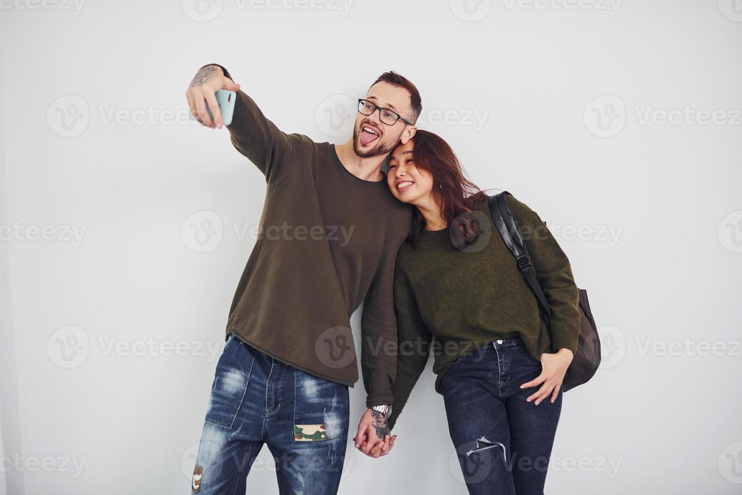 Fröhliches multiethnisches Paar in Freizeitkleidung, das im Studio vor weißem Hintergrund ein Selfie macht foto