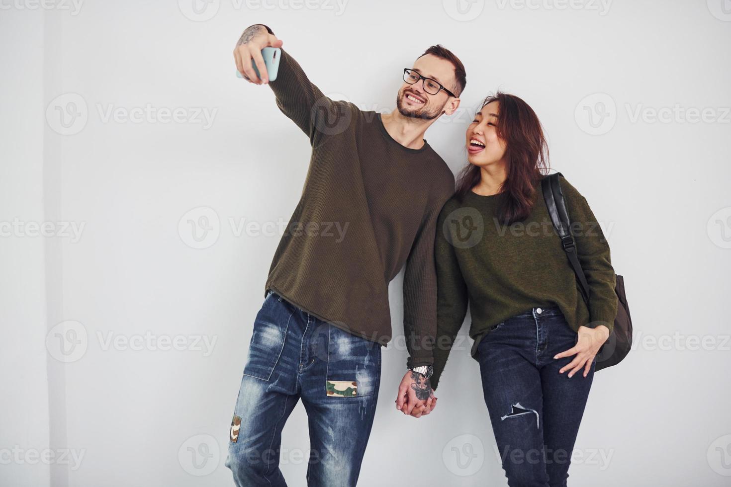 Fröhliches multiethnisches Paar in Freizeitkleidung, das im Studio vor weißem Hintergrund ein Selfie macht foto