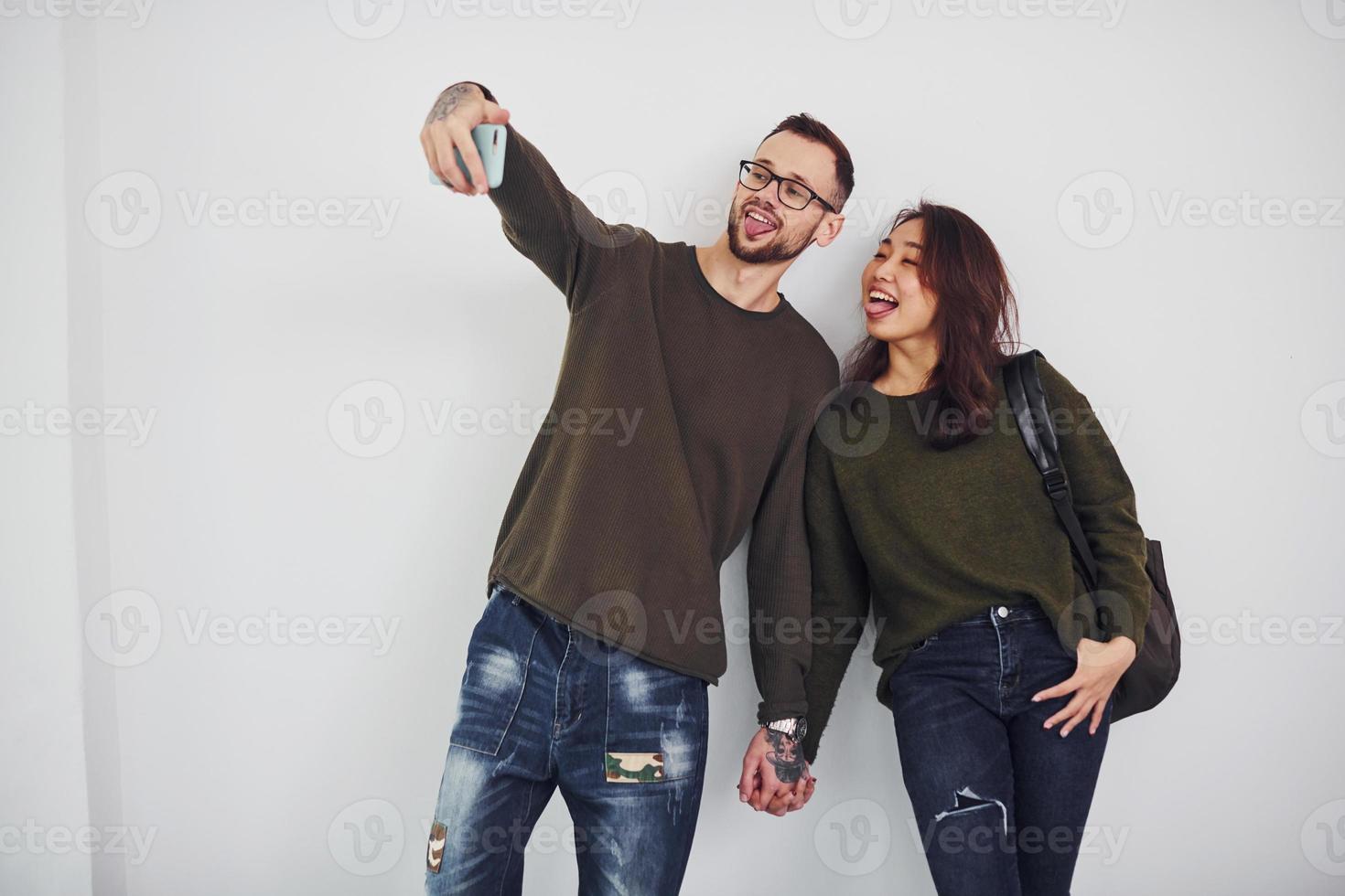 Fröhliches multiethnisches Paar in Freizeitkleidung, das im Studio vor weißem Hintergrund ein Selfie macht foto