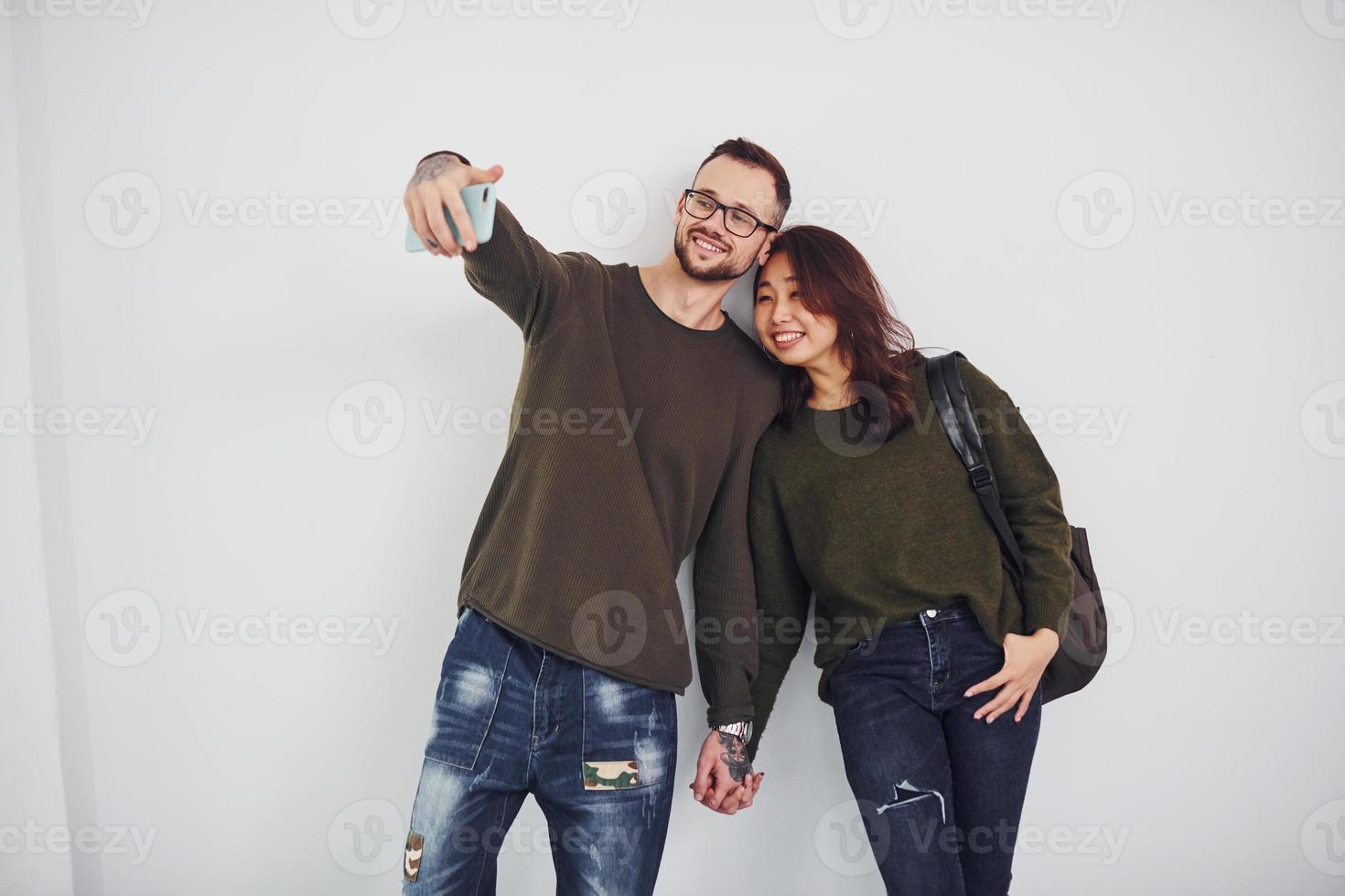 Fröhliches multiethnisches Paar in Freizeitkleidung, das im Studio vor weißem Hintergrund ein Selfie macht foto