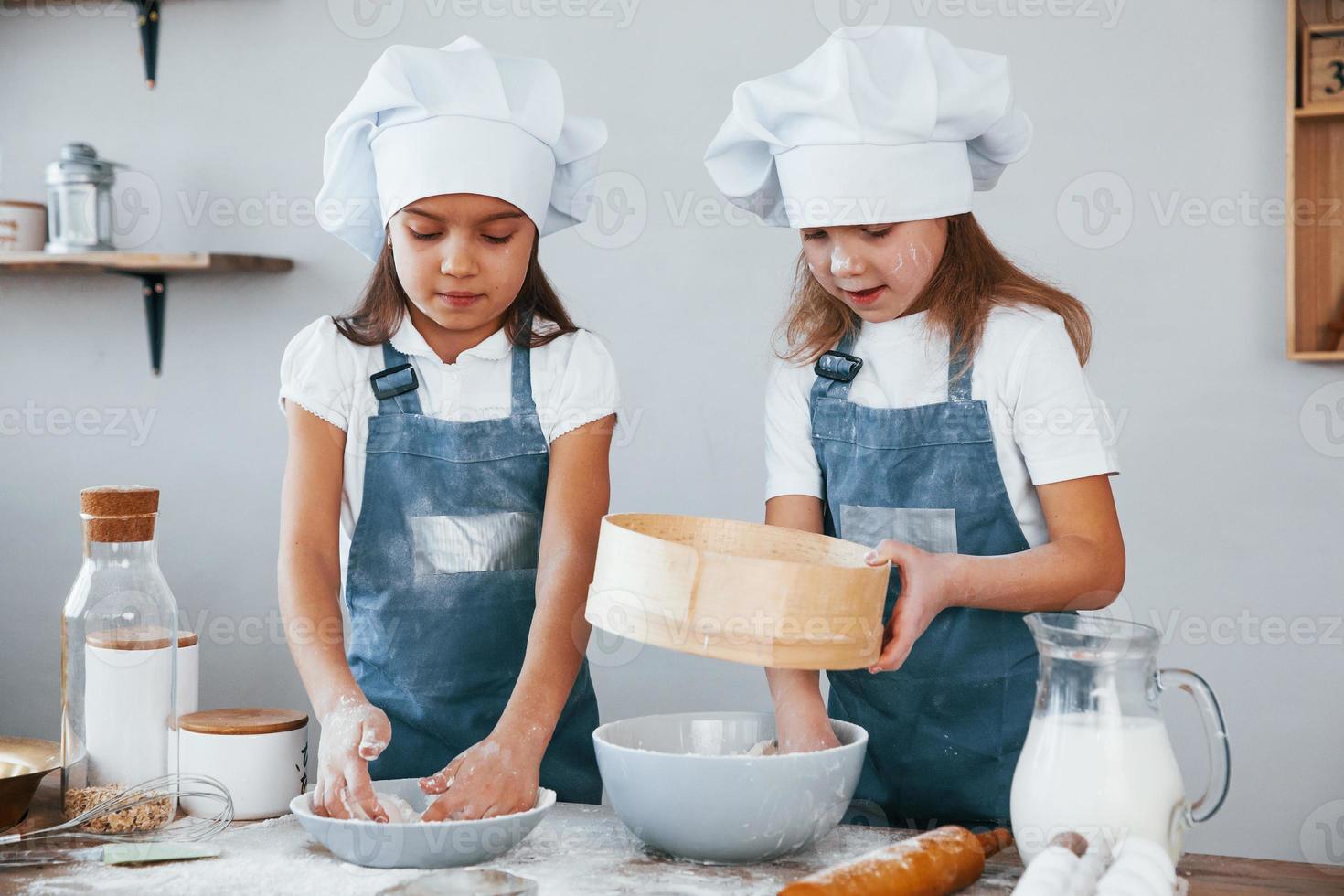 Zwei kleine Mädchen in blauer Kochuniform, die mit Mehl arbeiten, indem sie ein Sieb in der Küche verwenden foto