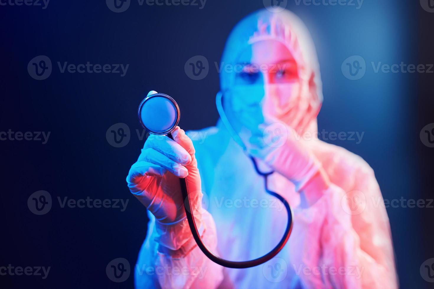 krankenschwester in maske und weißer uniform und mit stethoskop, das in einem neonbeleuchteten raum steht foto