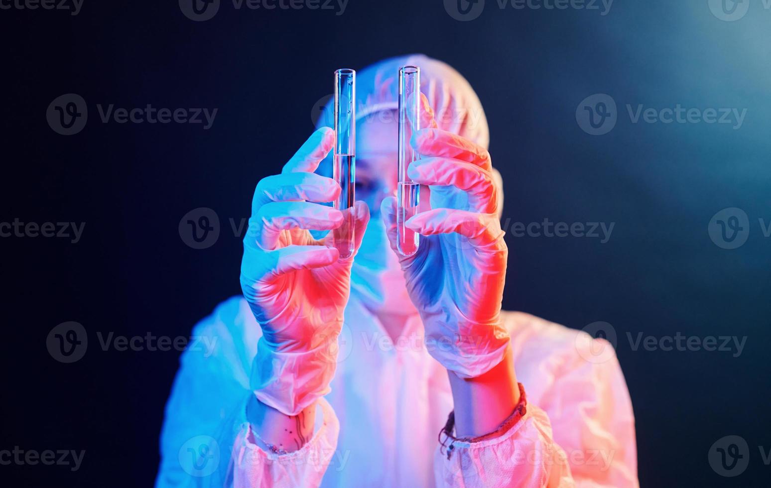 krankenschwester in maske und weißer uniform, die in einem neonbeleuchteten raum steht und röhrchen mit proben hält foto