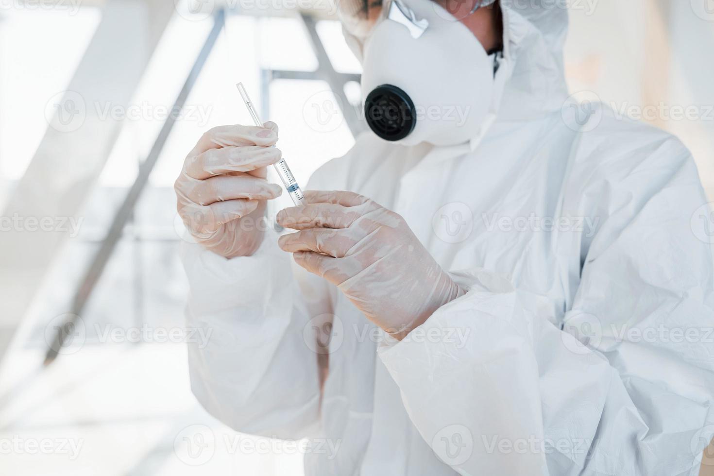 hält Spritze mit Medizin. Ärztin in Laborkittel, Schutzbrille und Maske im Innenbereich foto