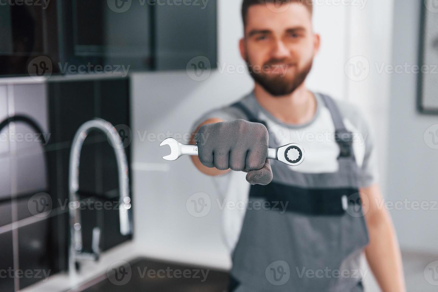 Nahaufnahme eines jungen professionellen Klempners in grauer Uniform, der einen Schraubenschlüssel in der Hand in der Küche hält foto