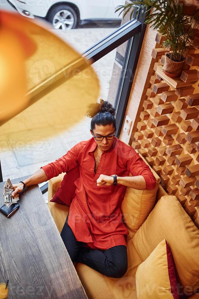 Blick von oben auf den reichen Mann in roter und schwarzer Kleidung, der mit viel Geld in der Hand drinnen im Café sitzt foto