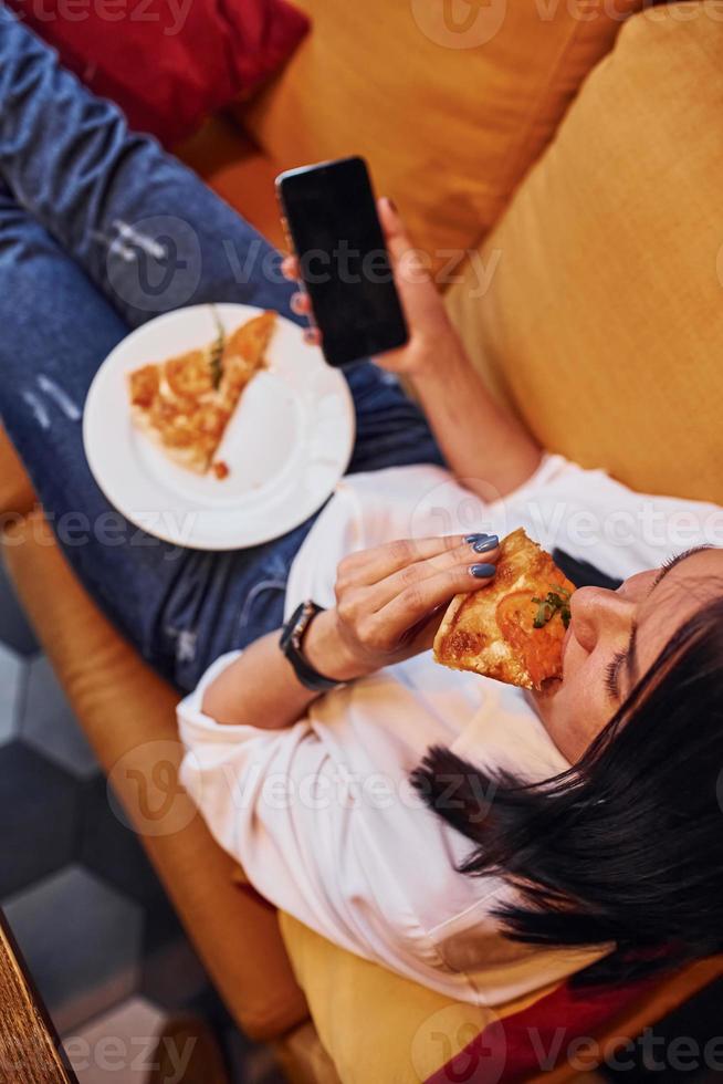 Junge Brünette, die drinnen mit Pizza und Smartphone in der Hand sitzt foto