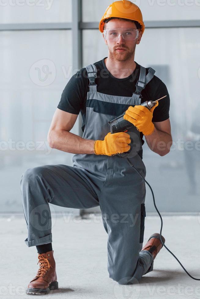 Mann in grauer Uniform mit Bohrer in der Hand, der tagsüber drinnen in einem modernen großen Büro steht foto