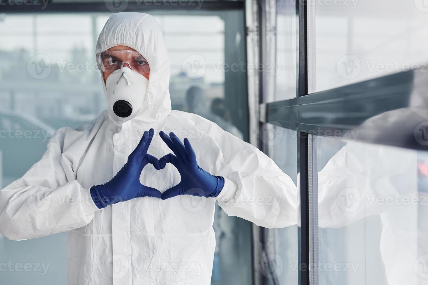 männlicher arztwissenschaftler in laborkittel, defensiver brille und maske zeigt liebesgeste mit den händen foto