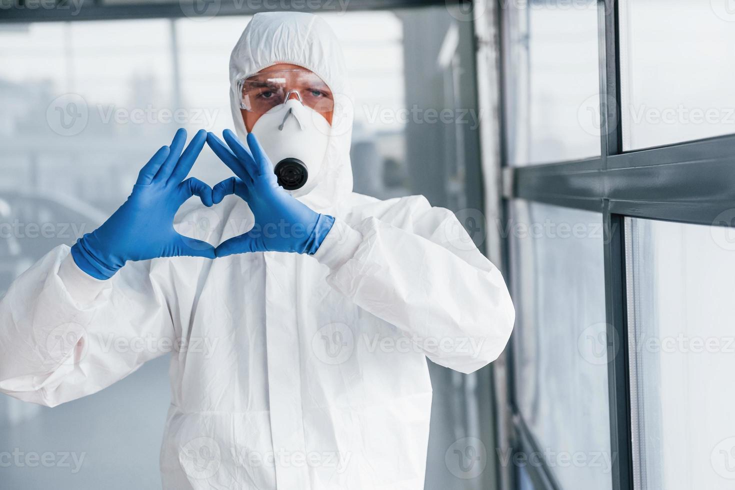männlicher arztwissenschaftler in laborkittel, defensiver brille und maske zeigt liebesgeste mit den händen foto