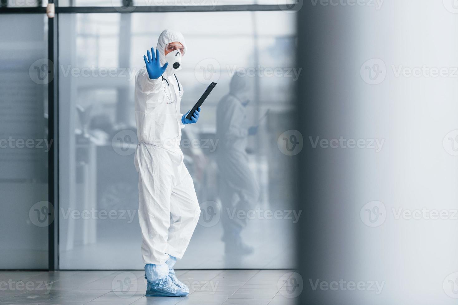 zeigt Stop-Geste von Hand. männlicher arztwissenschaftler in laborkittel, defensiver brille und maske, die notizblock in den händen hält foto