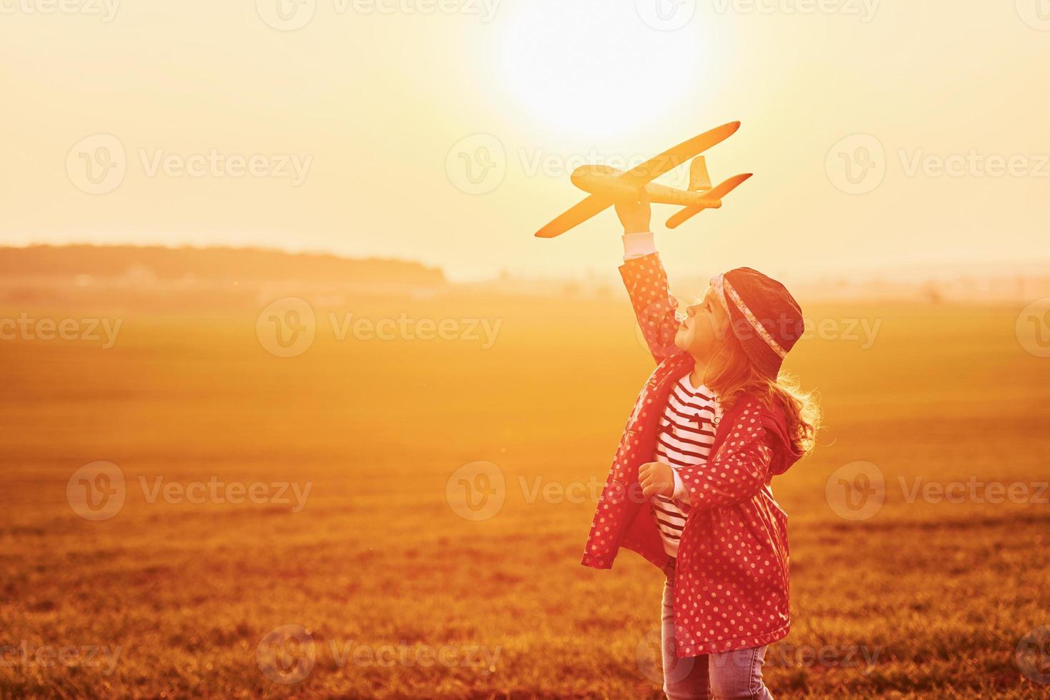 von orangefarbenem Sonnenlicht beleuchtet. süßes kleines mädchen hat tagsüber spaß mit spielzeugflugzeug auf dem schönen feld foto