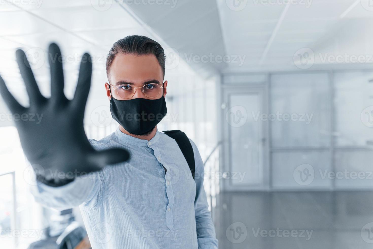 Stoppgeste von Hand zeigen. in Schutzmaske und Handschuhen. junger gutaussehender mann in formeller kleidung drinnen im büro tagsüber foto