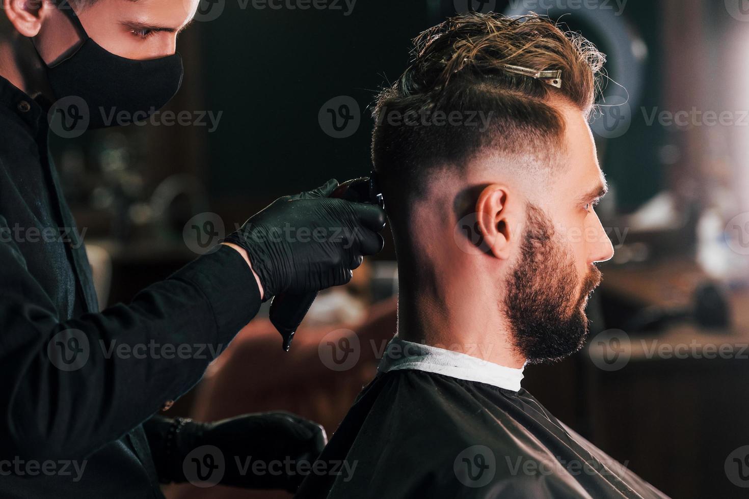 junger bärtiger mann, der im friseursalon sitzt und sich von einem kerl in schwarzer schutzmaske die haare schneiden lässt foto