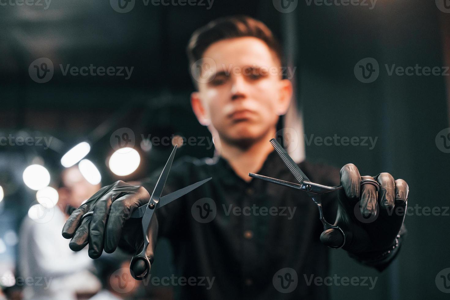 Porträt eines jungen Mannes in schwarzer Kleidung, der drinnen im Friseursalon steht und eine Schere hält foto