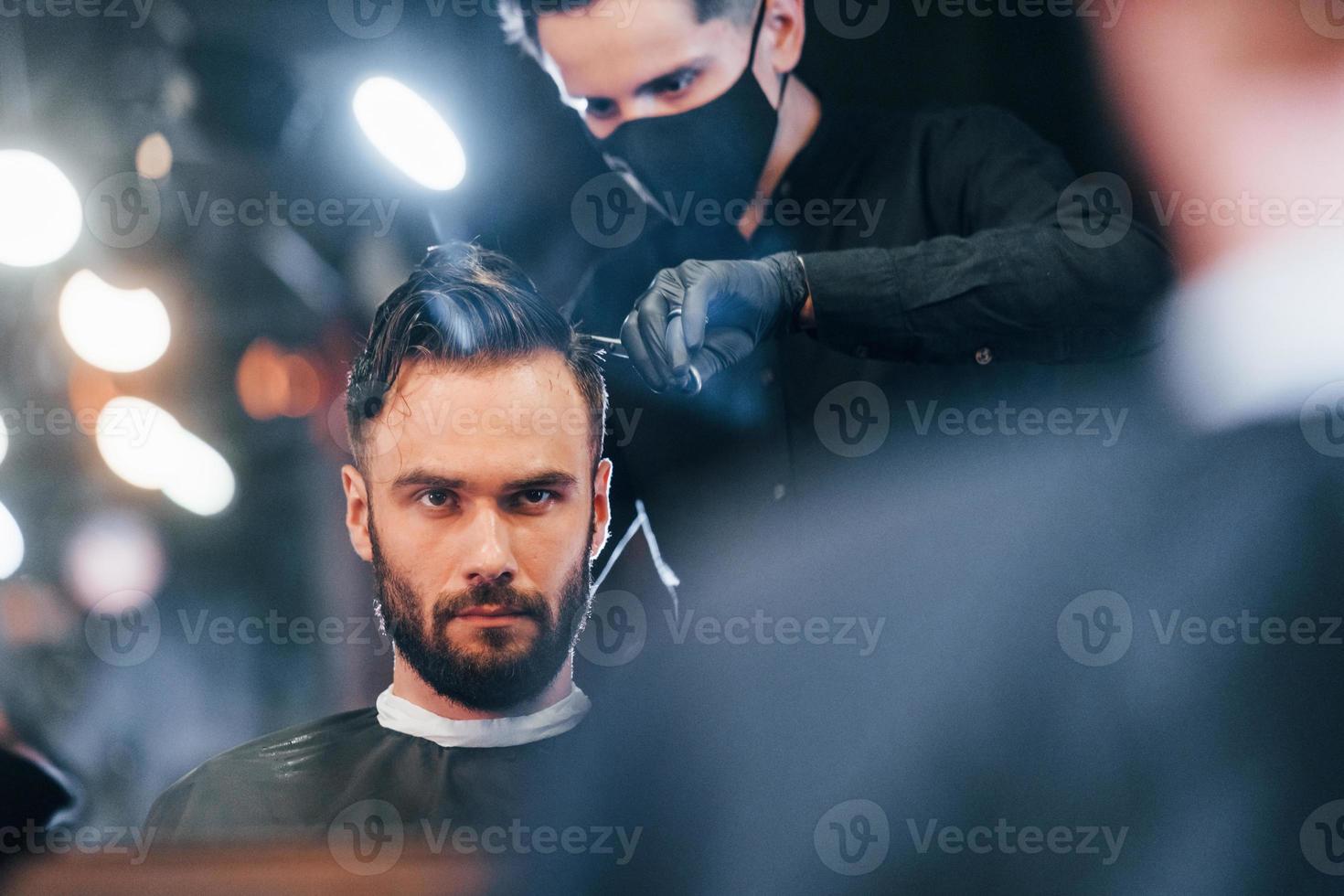 Vorderansicht des jungen bärtigen Mannes, der im Friseursalon von einem Mann in schwarzer Schutzmaske sitzt und sich die Haare schneiden lässt foto