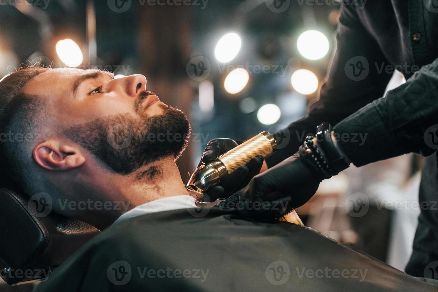 Junger Mann mit stylischer Frisur sitzt und lässt sich im Friseurladen den Bart rasieren foto