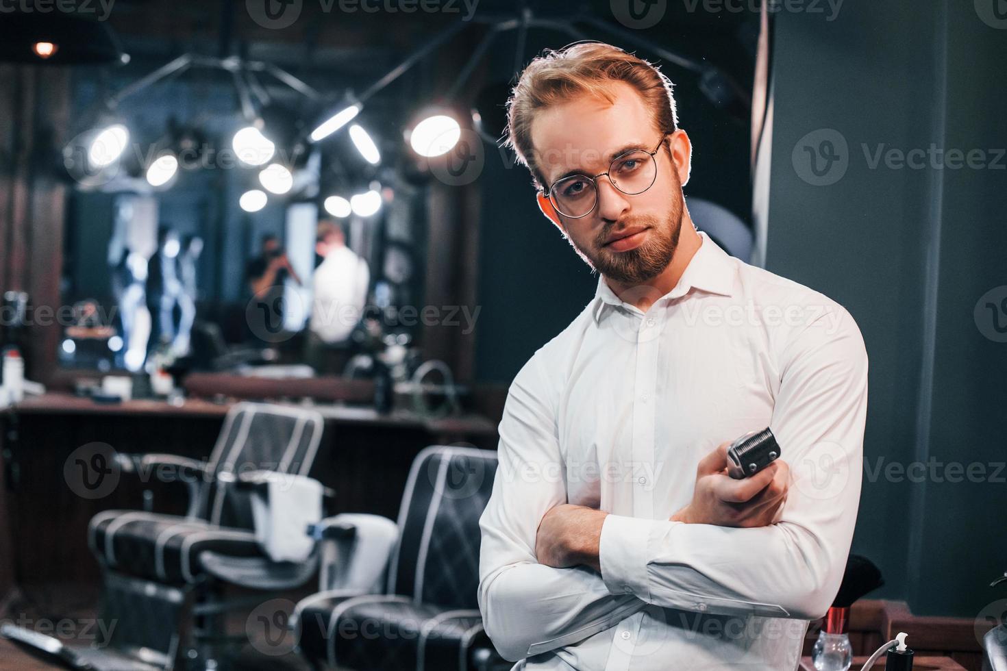 Porträt eines jungen Mannes in weißer Kleidung, der drinnen im Friseursalon steht und eine Haarschneidemaschine hält foto