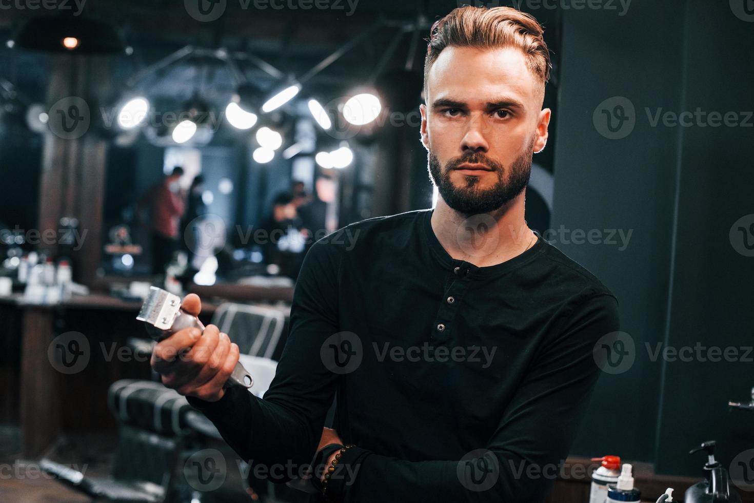 Junger bärtiger Mann, der im Friseursalon steht und eine Haarschneidemaschine hält foto