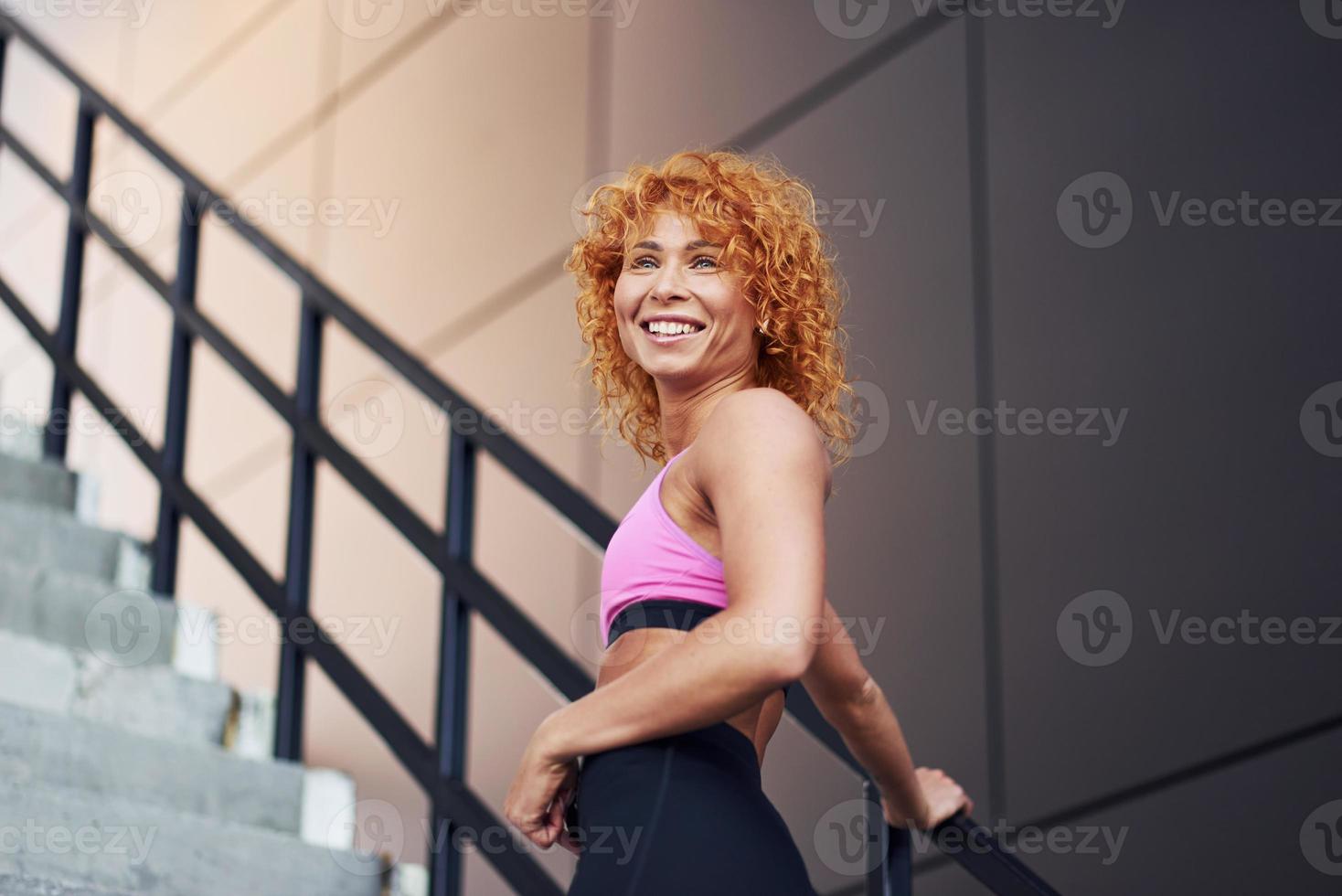 junge europäische rothaarige frau in sportlicher kleidung, die ihren körper zeigt und nach fitnessübungen im freien posiert foto