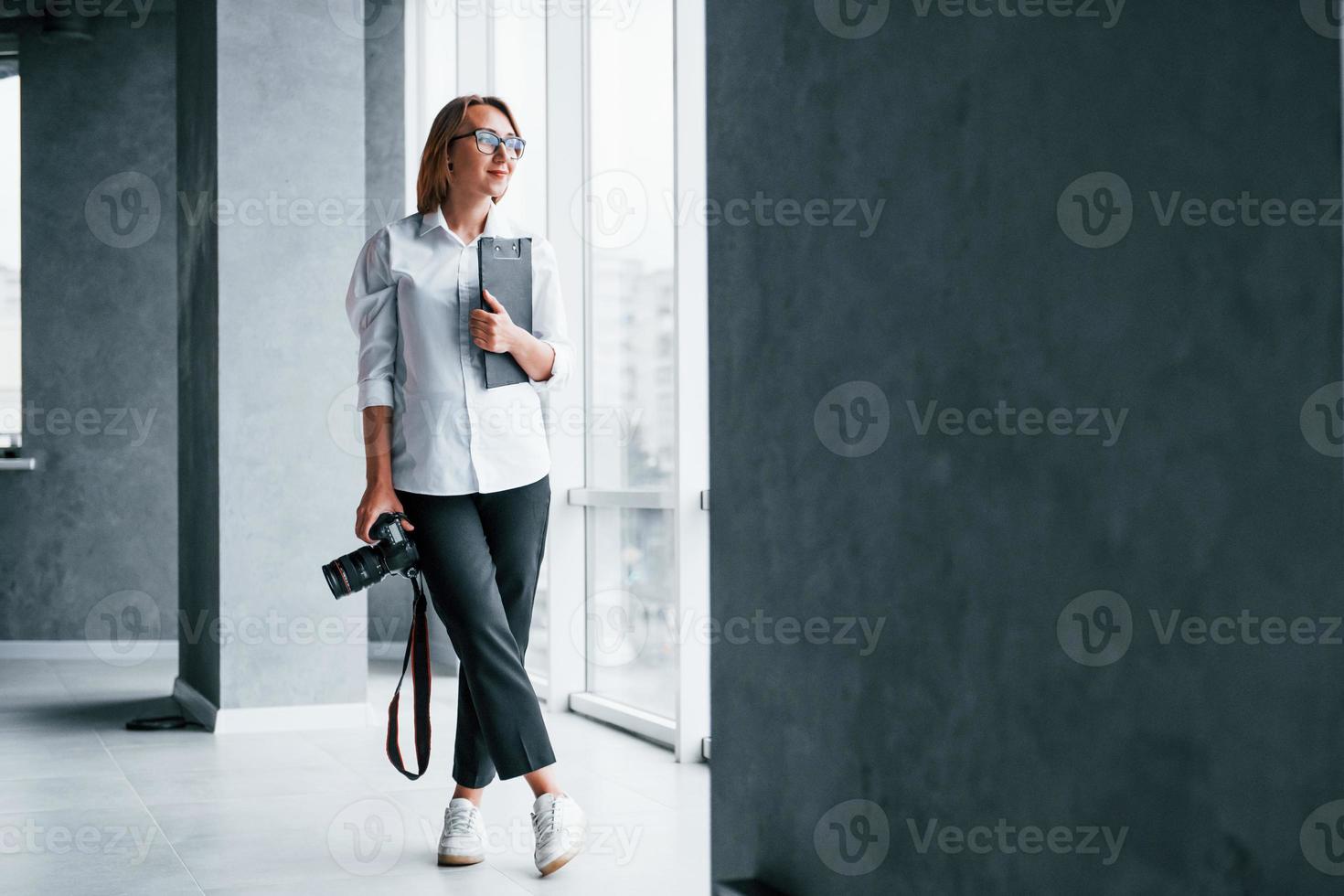 Frau in formeller Kleidung und mit Kamera in der Hand, die in einem leeren Raum steht foto