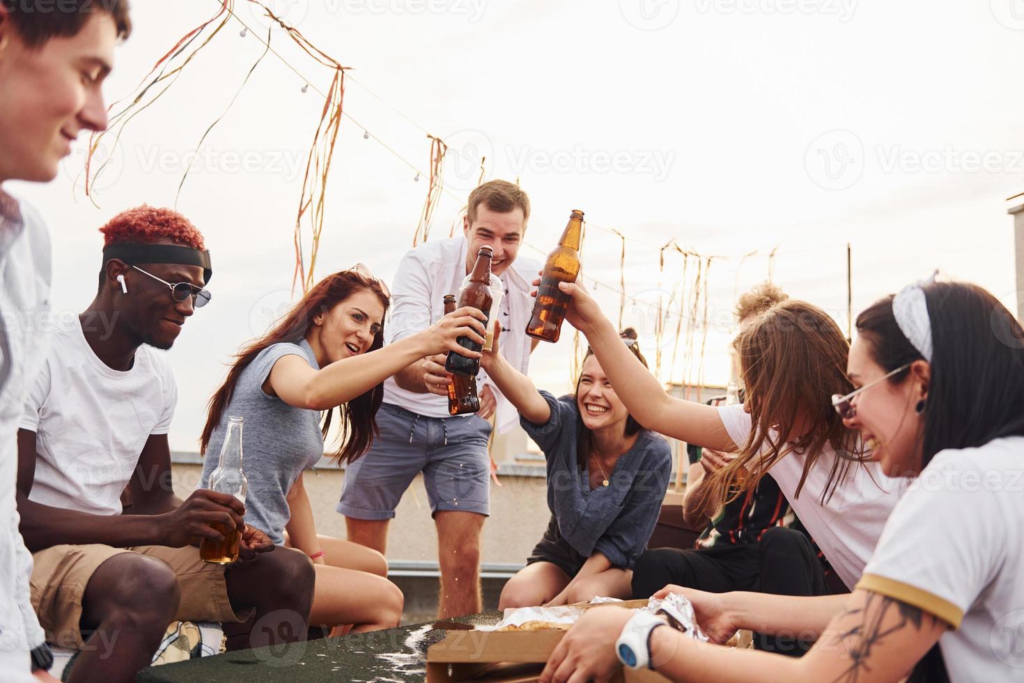 Anfeuern durch Flaschen mit Bier. eine gruppe junger leute in lässiger kleidung feiert tagsüber zusammen eine party auf dem dach foto