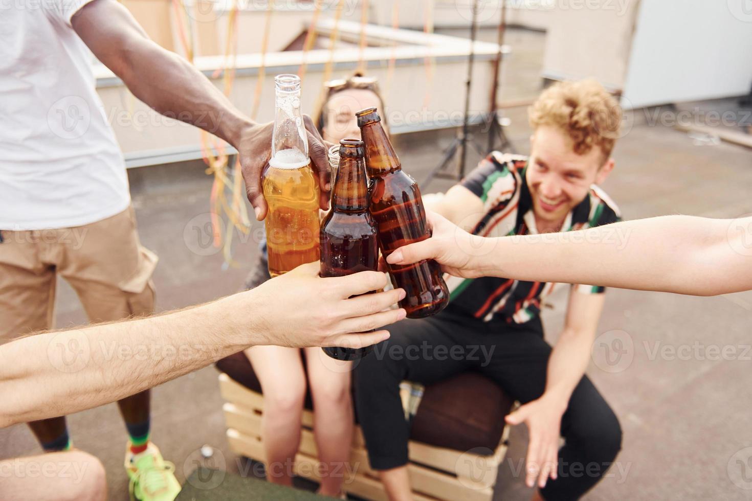 Anfeuern durch Flaschen mit Bier. eine gruppe junger leute in lässiger kleidung feiert tagsüber zusammen eine party auf dem dach foto