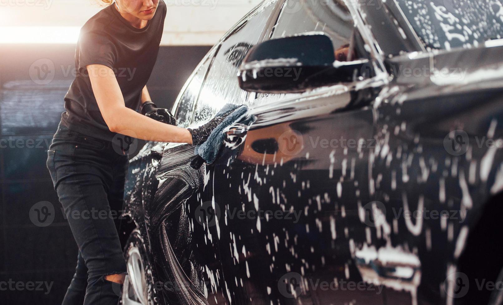 wischt Fahrzeug, das in weißer Seife ist. Modernes schwarzes Auto wird von einer Frau in der Autowaschanlage gereinigt foto