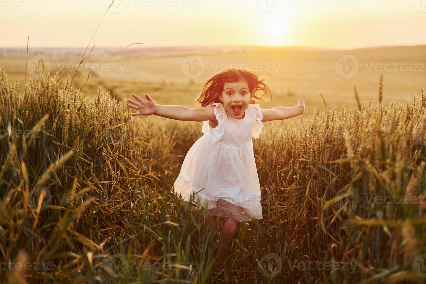 Fröhliches kleines Mädchen in weißem Kleid, das zur Sommertageszeit auf dem landwirtschaftlichen Gebiet läuft foto