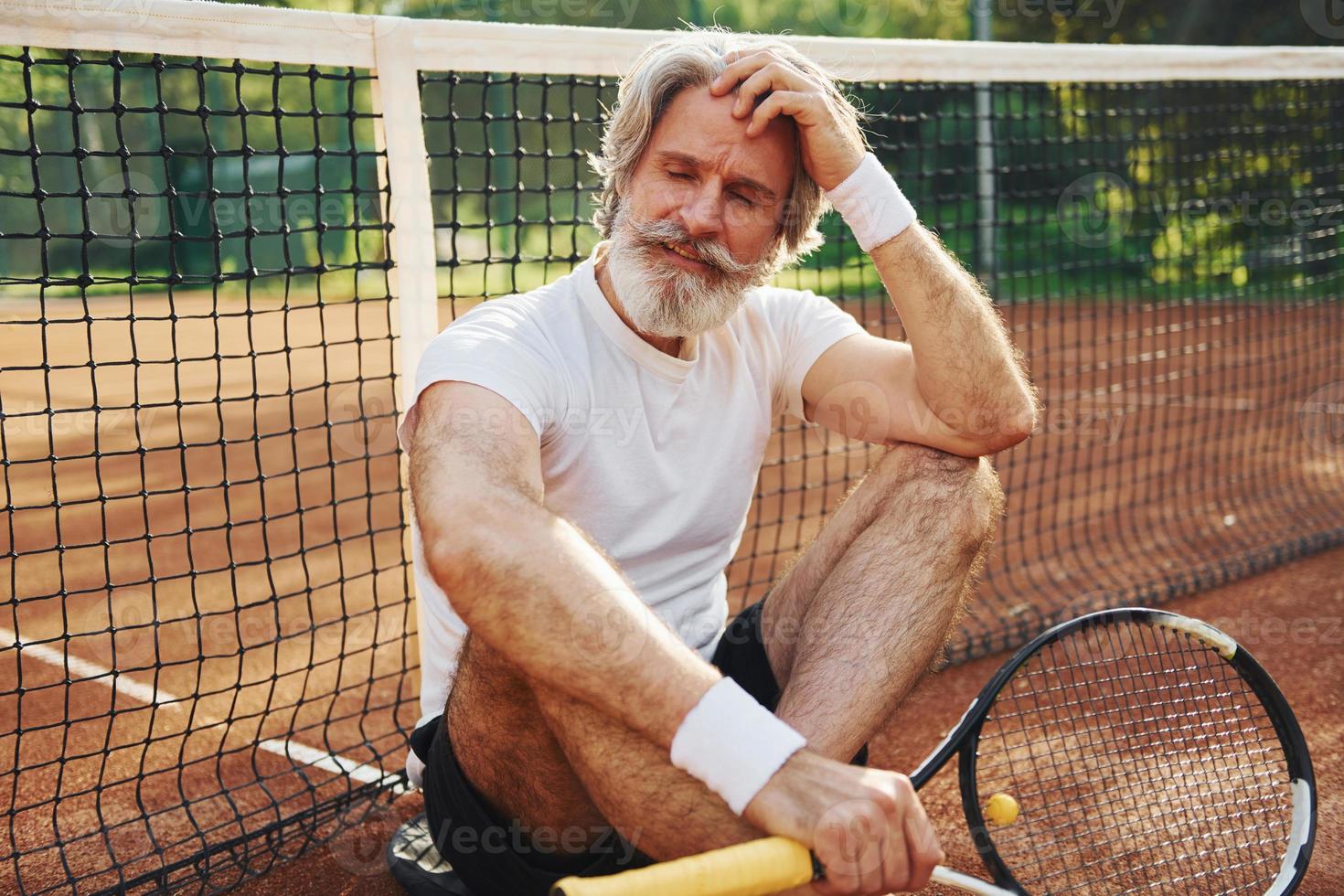 sitzt auf dem Boden und macht Pause. Älterer, moderner, stilvoller Mann mit Schläger im Freien auf dem Tennisplatz tagsüber foto