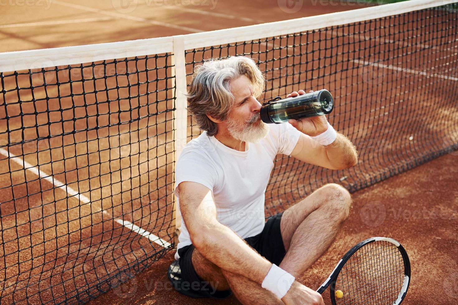 Wasser trinken und auf dem Boden sitzen. Älterer, moderner, stilvoller Mann mit Schläger im Freien auf dem Tennisplatz tagsüber foto