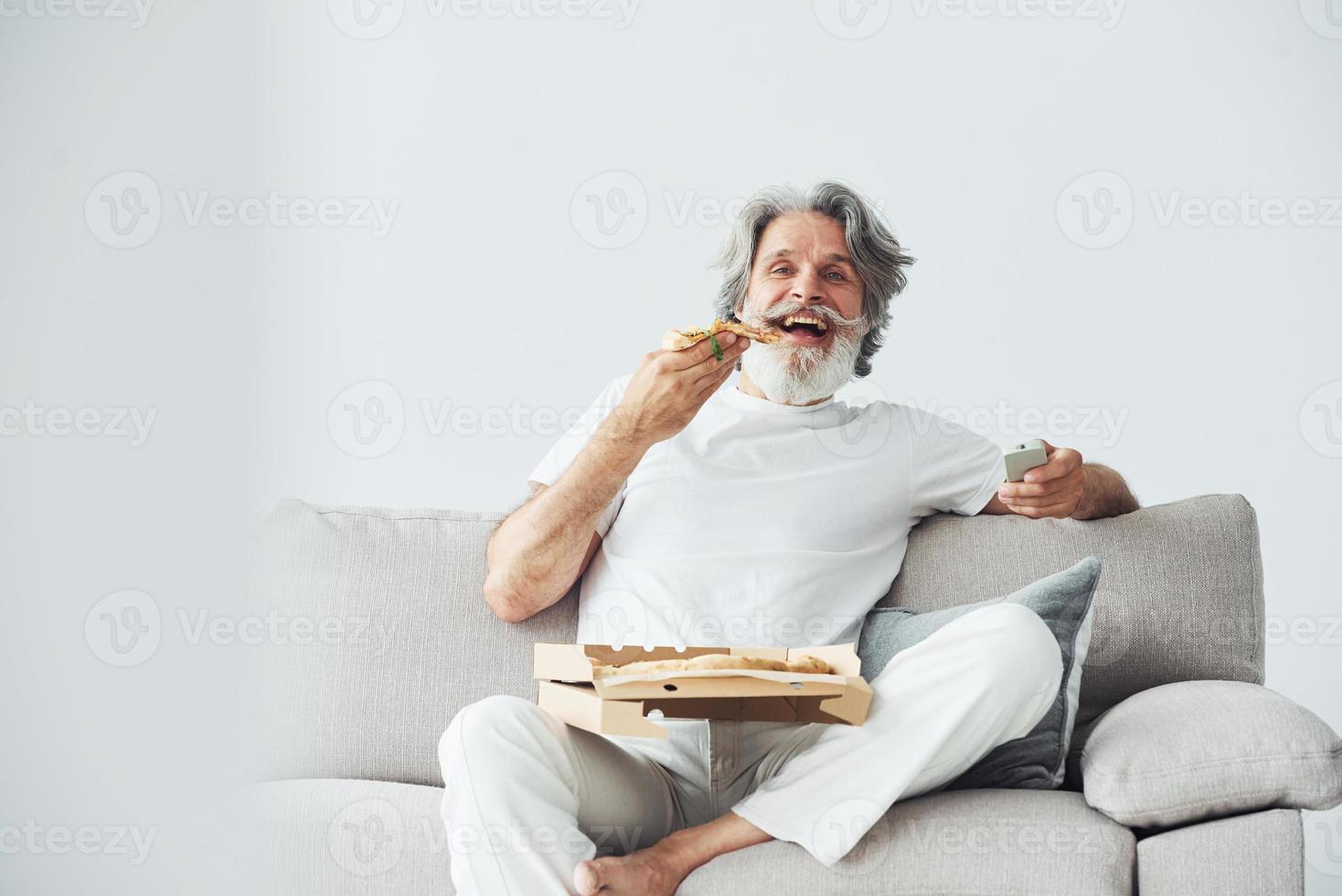 mit leckerer Pizza. schaut sich eine Fernsehsendung an. älterer stilvoller moderner mann mit grauem haar und bart zuhause foto