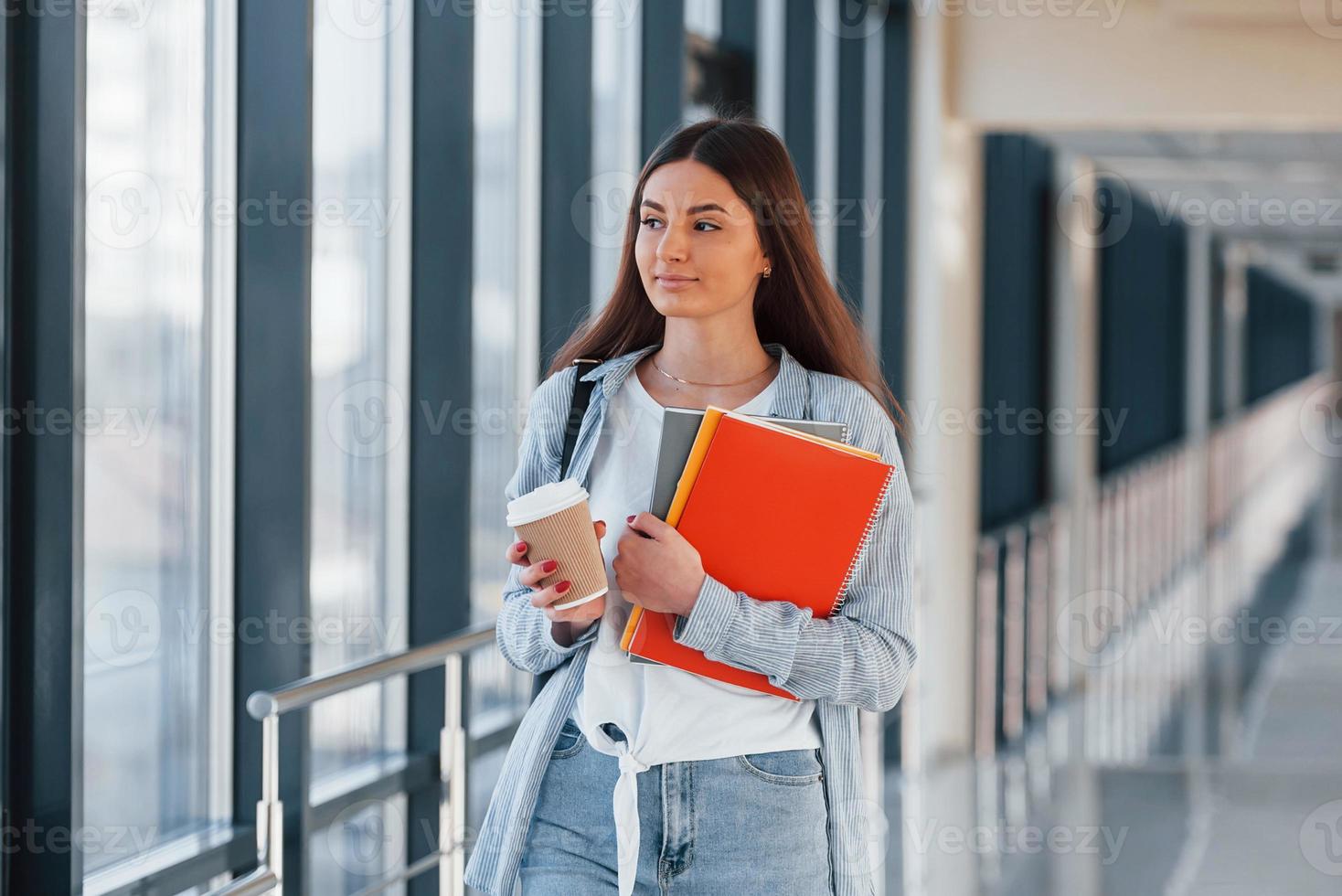 junge studentin ist im flur eines colleges und hält notizblöcke und eine tasse getränk foto