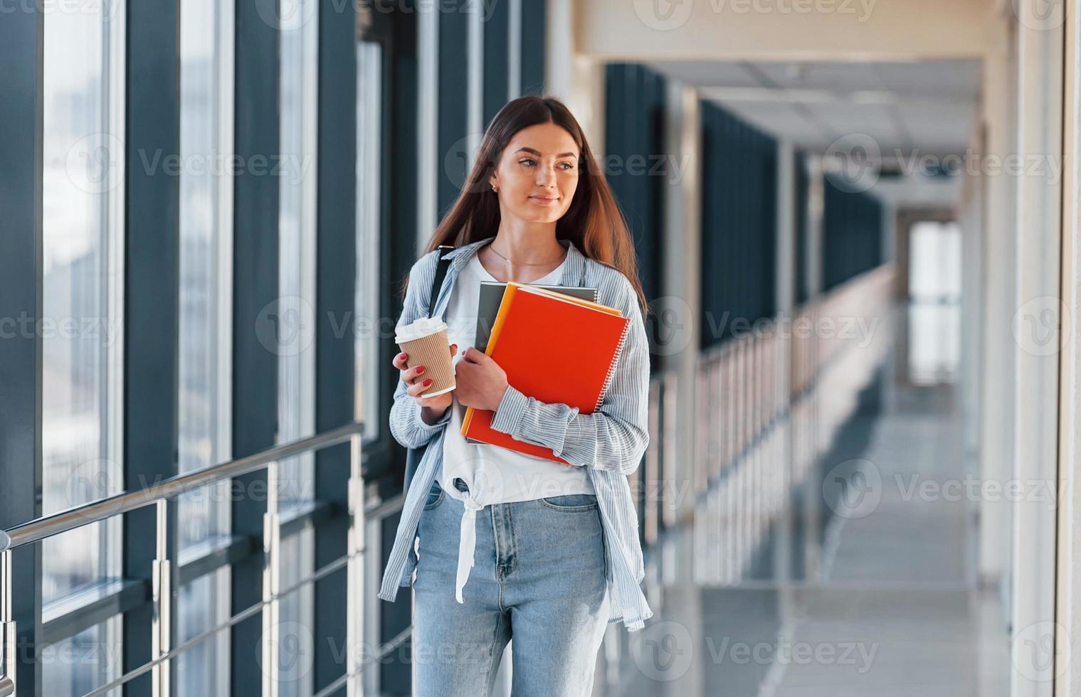 junge studentin ist im flur eines colleges und hält notizblöcke und eine tasse getränk foto