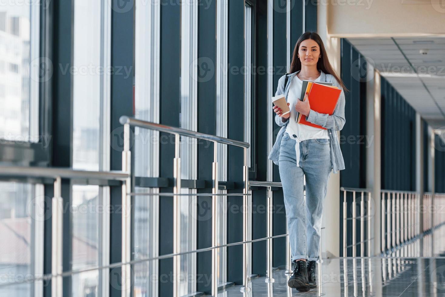 junge studentin ist im flur eines colleges und hält notizblöcke und eine tasse getränk foto