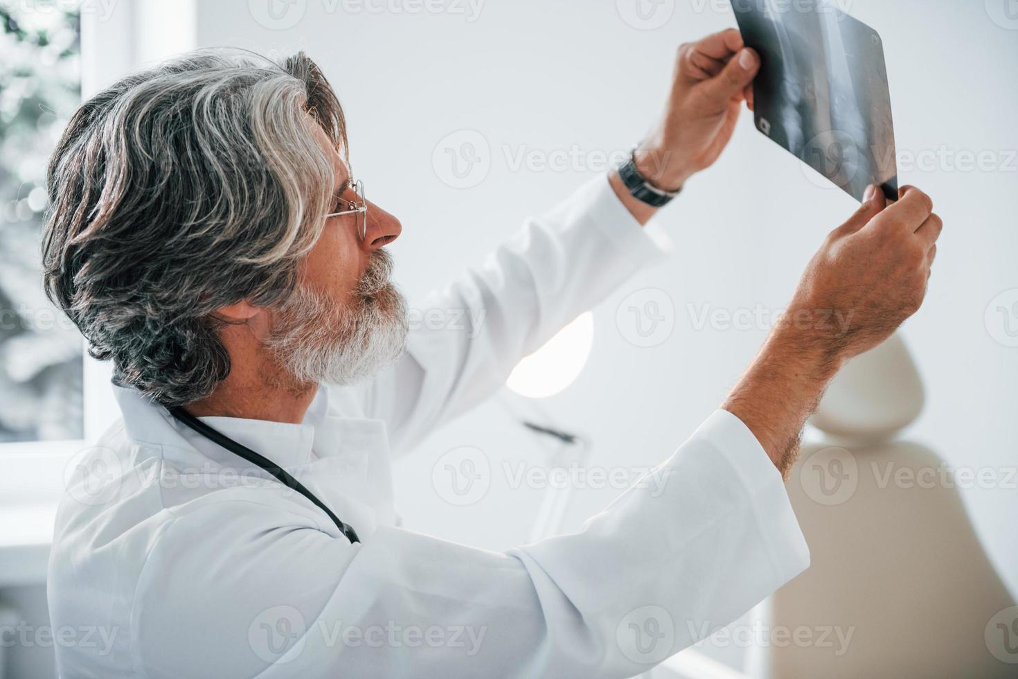 sieht sich das Röntgenbild an. leitender männlicher arzt mit grauem haar und bart im weißen kittel ist drinnen in der klinik foto