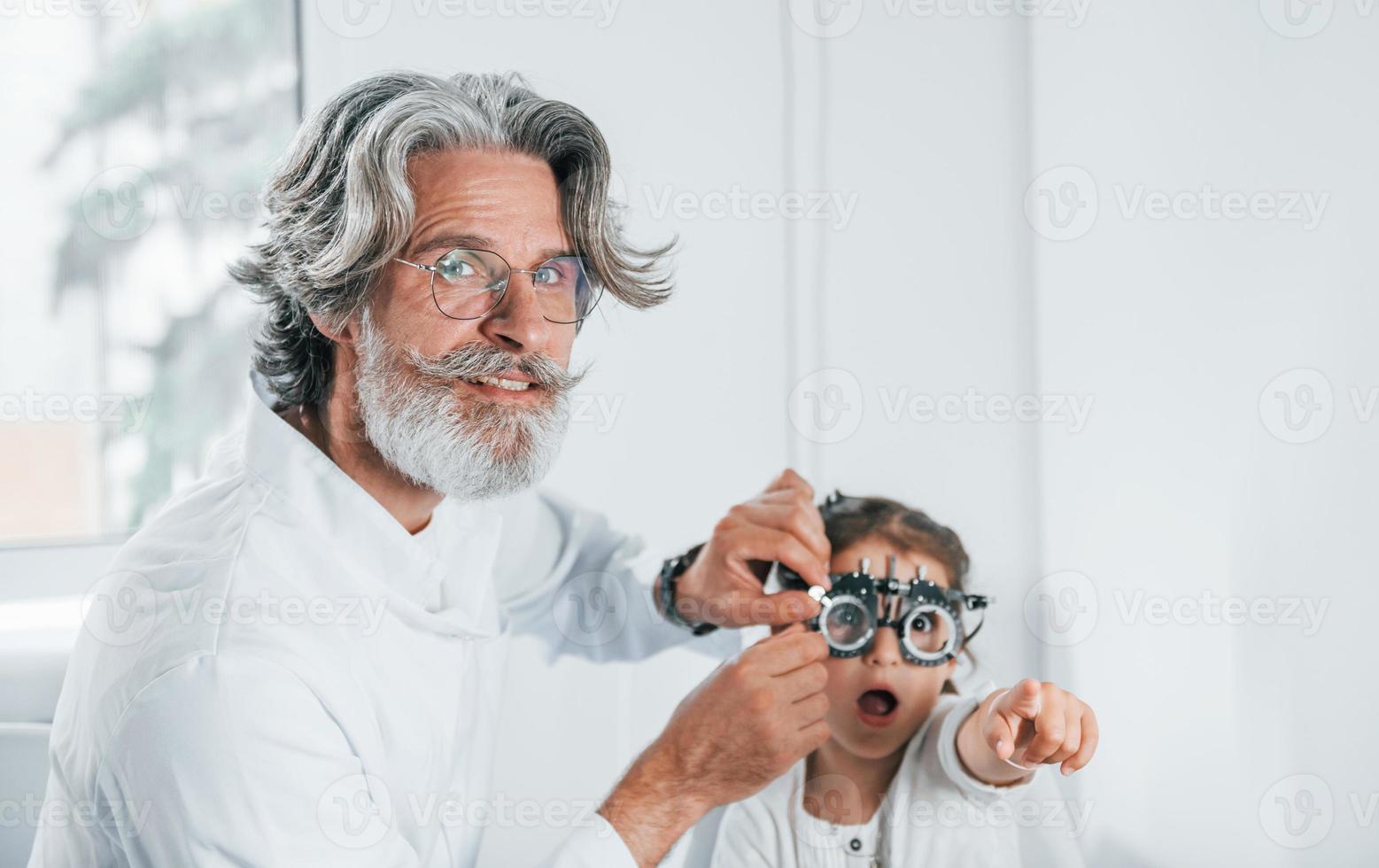 leitender männlicher Augenarzt mit grauem Haar und Bart im weißen Kittel ist drinnen in der Klinik und testet den Anblick eines kleinen Mädchens foto