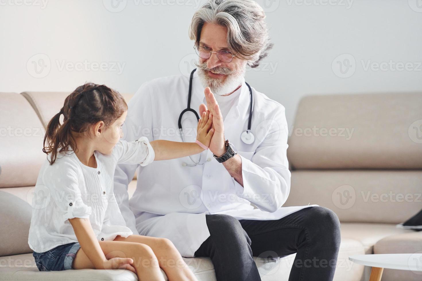 gibt dem kleinen Mädchen High Five. leitender männlicher arzt mit grauem haar und bart im weißen kittel ist drinnen in der klinik foto