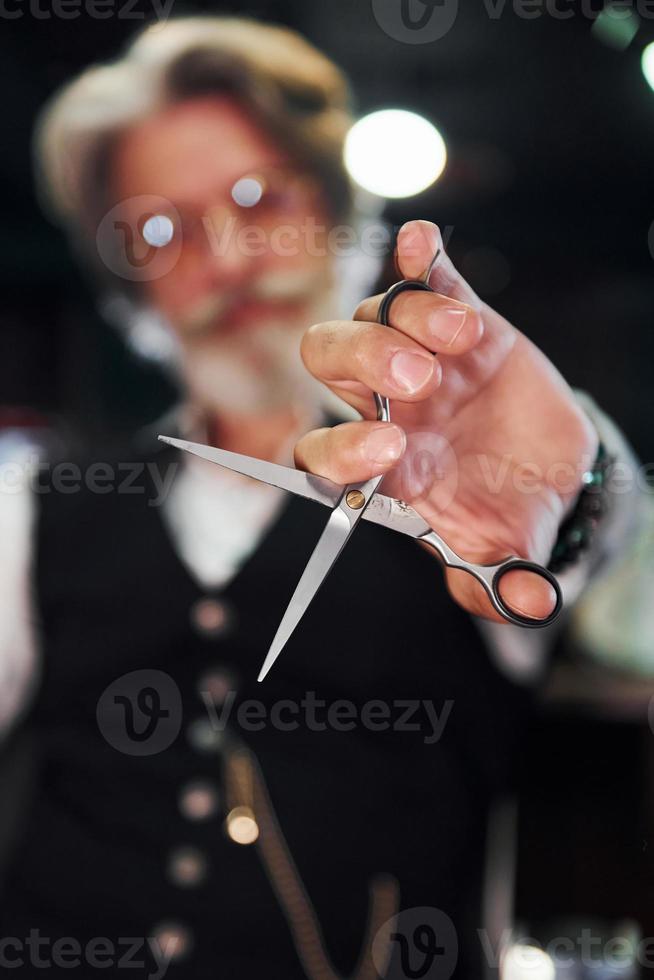 sitzt auf dem stuhl im friseursalon. Stilvoller moderner Senior mit grauem Haar und Bart ist drinnen foto