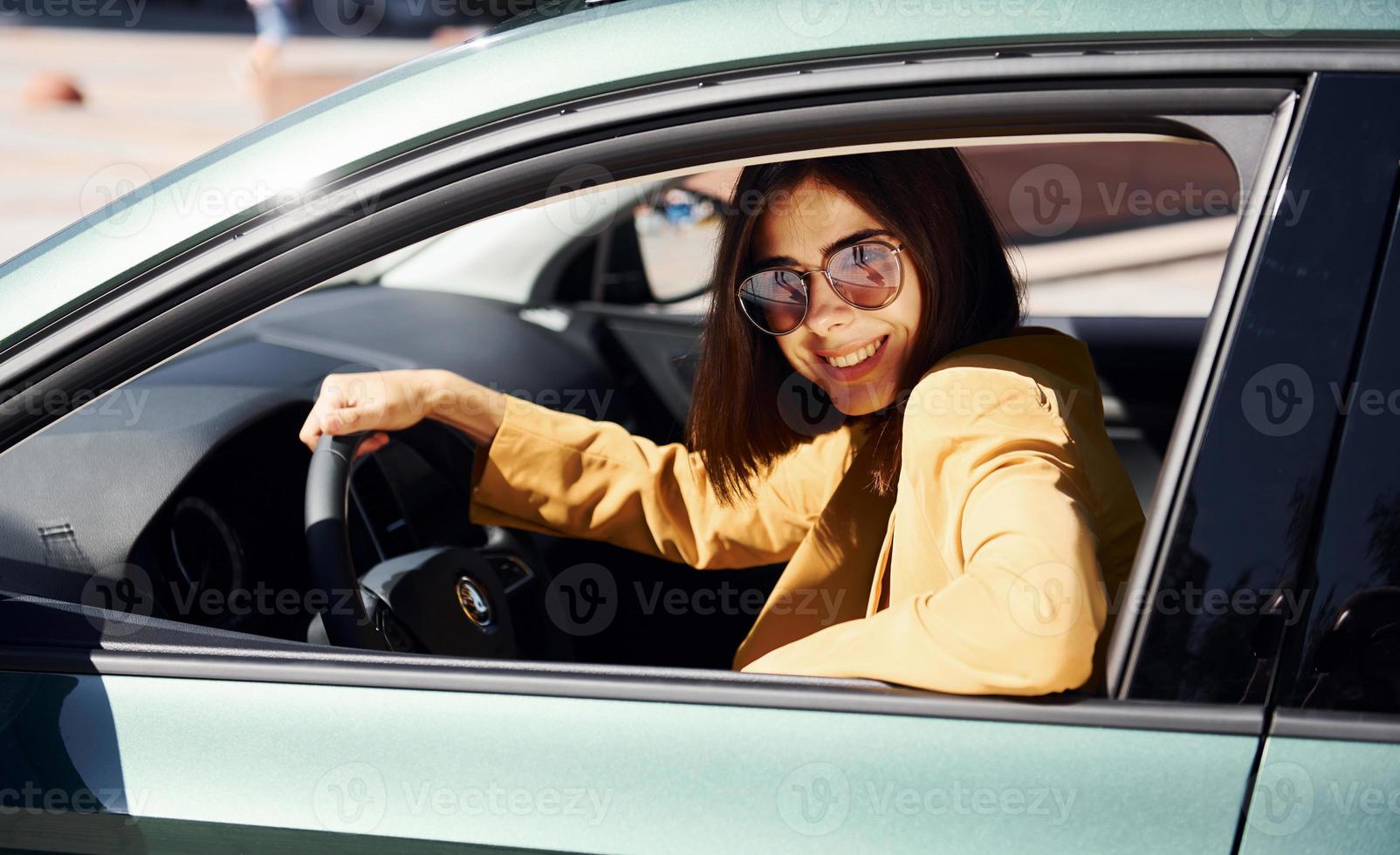 Glücklicher Autobesitzer sitzt im Fahrzeug. junge modische frau in burgunderfarbenem mantel tagsüber mit ihrem auto foto