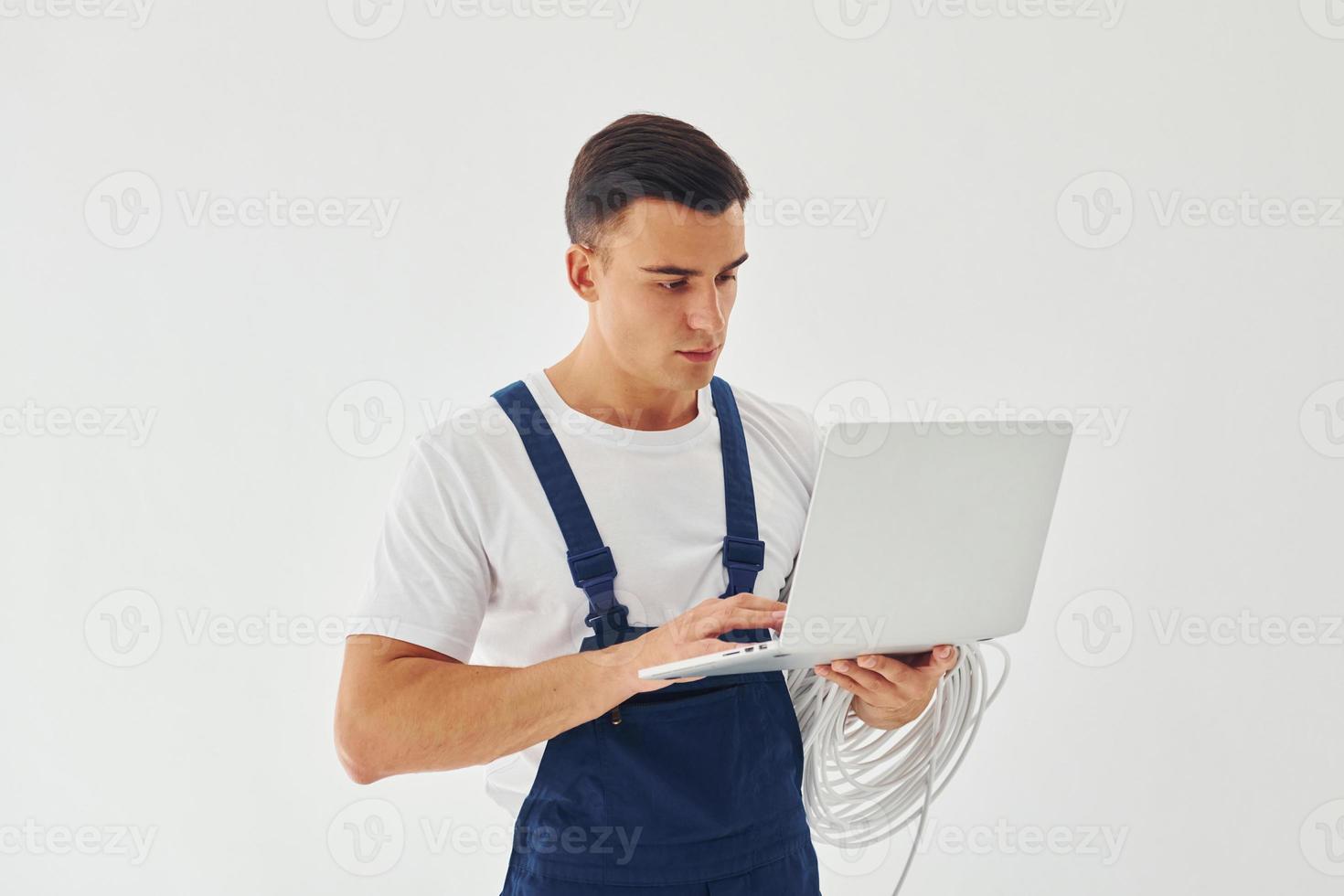 nutzt Laptop. männlicher Arbeiter in blauer Uniform, der im Studio vor weißem Hintergrund steht foto