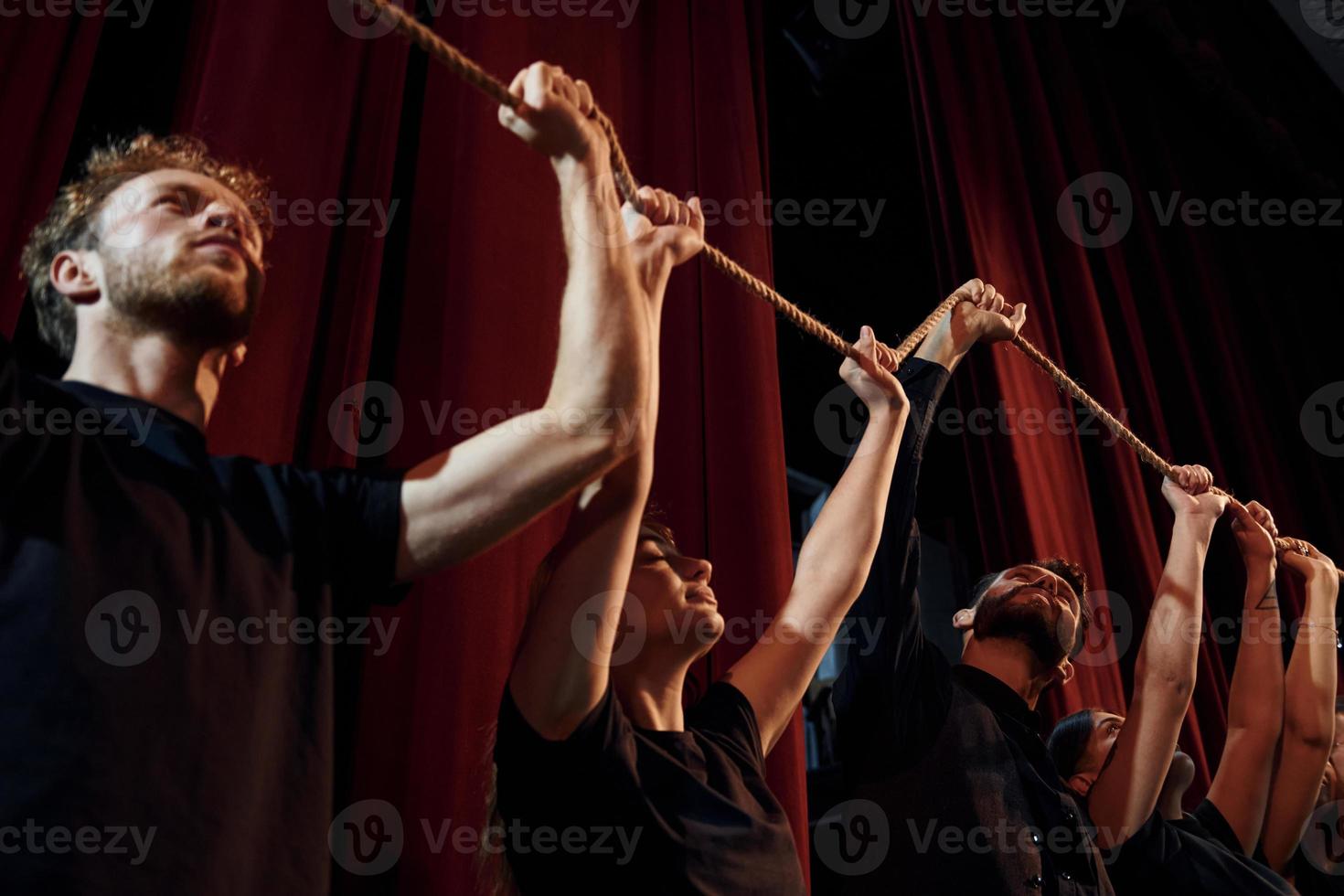 Seil in den Händen über den Köpfen halten. Gruppe von Schauspielern in dunkler Kleidung bei der Probe im Theater foto