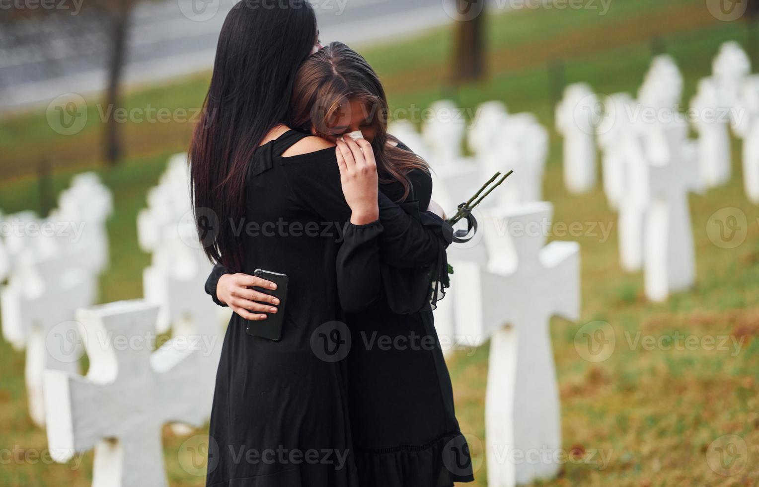 sich umarmen und weinen. Zwei junge Frauen in schwarzer Kleidung besuchen den Friedhof mit vielen weißen Kreuzen. Vorstellung von Begräbnis und Tod foto