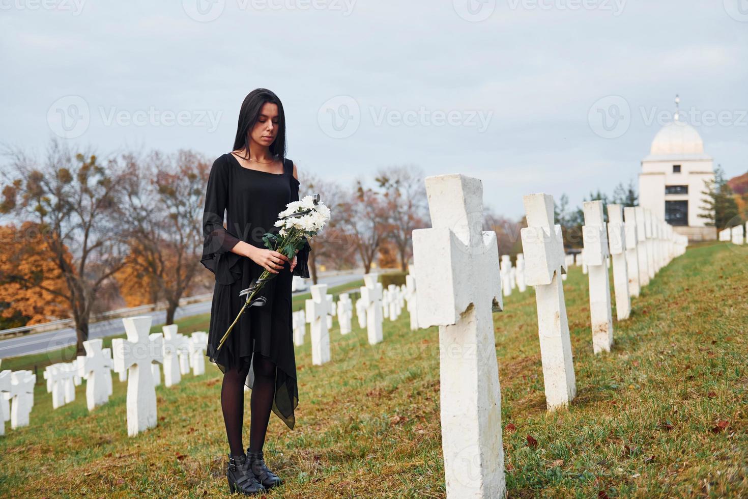 mit Blumen in den Händen. junge Frau in schwarzer Kleidung, die den Friedhof mit vielen weißen Kreuzen besucht. Vorstellung von Begräbnis und Tod foto