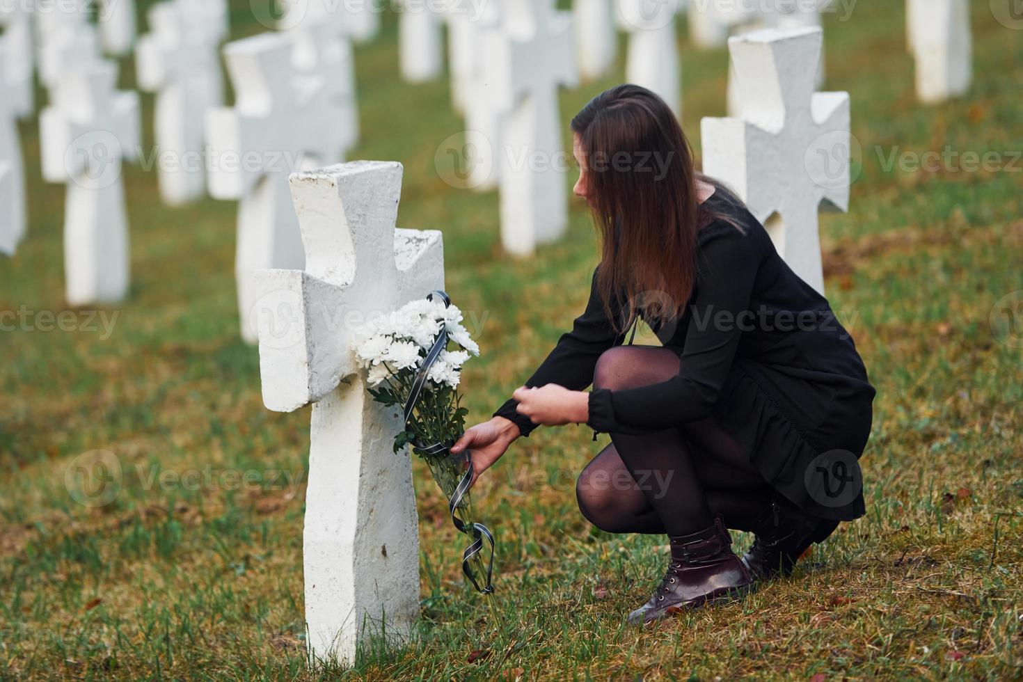 gibt Respekt, indem er Blumen legt. junge Frau in schwarzer Kleidung, die den Friedhof mit vielen weißen Kreuzen besucht. Vorstellung von Begräbnis und Tod foto