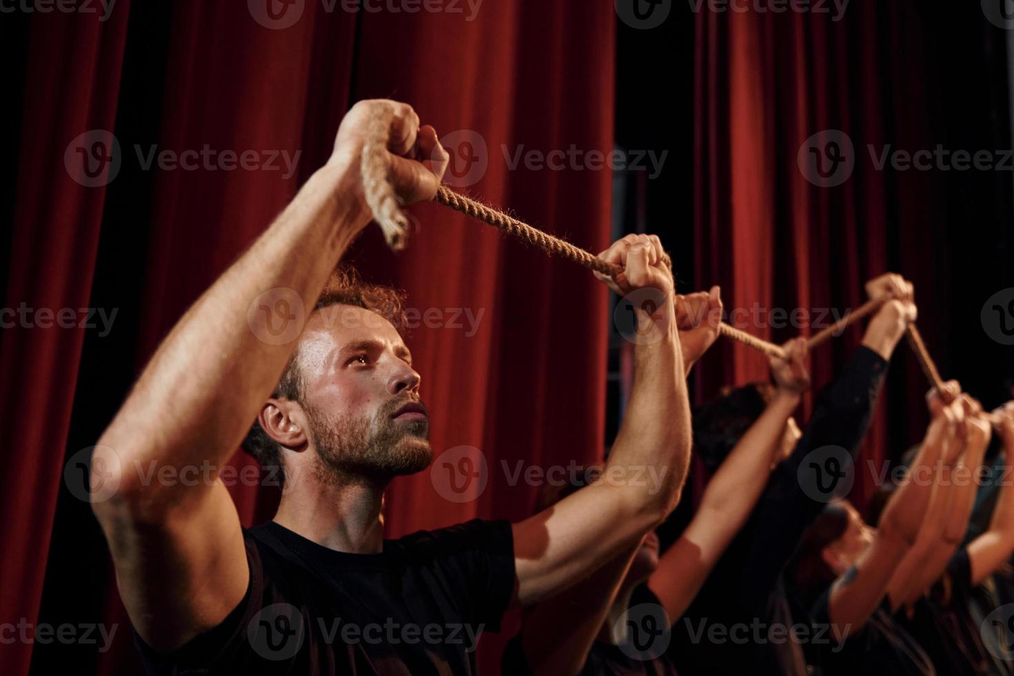 Seil in den Händen über den Köpfen halten. Gruppe von Schauspielern in dunkler Kleidung bei der Probe im Theater foto