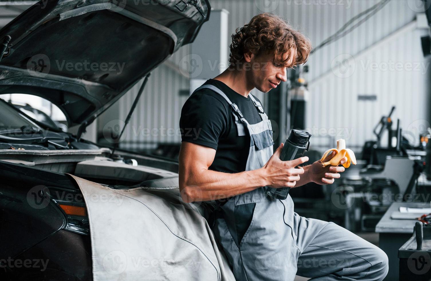 macht eine Pause. isst Bananen und trinkt Wasser. erwachsener mann in grauer uniform arbeitet im autosalon foto