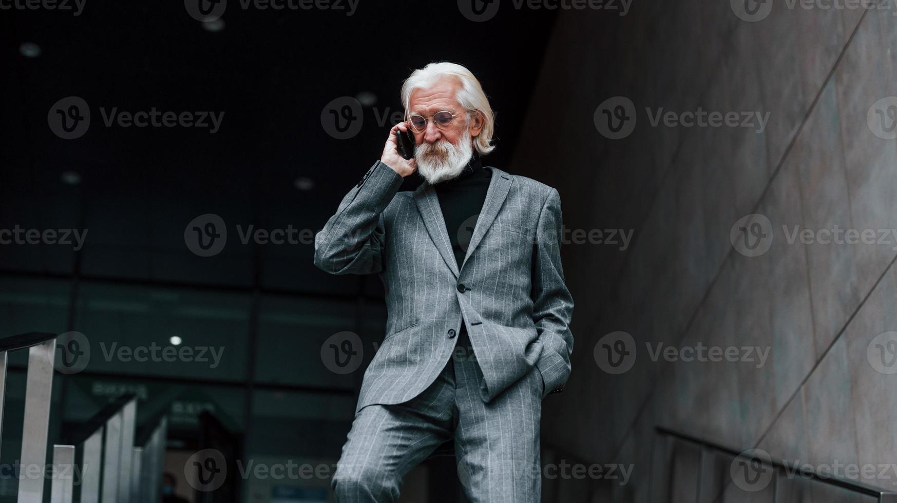 Konversation mit Telefon. Senior Geschäftsmann in formeller Kleidung, mit grauem Haar und Bart ist im Freien foto