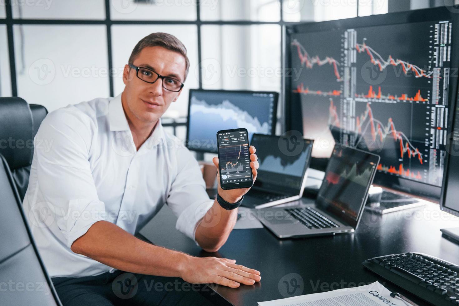 hält Telefon. junger männlicher börsenmakler arbeitet drinnen im büro. viele Displays foto